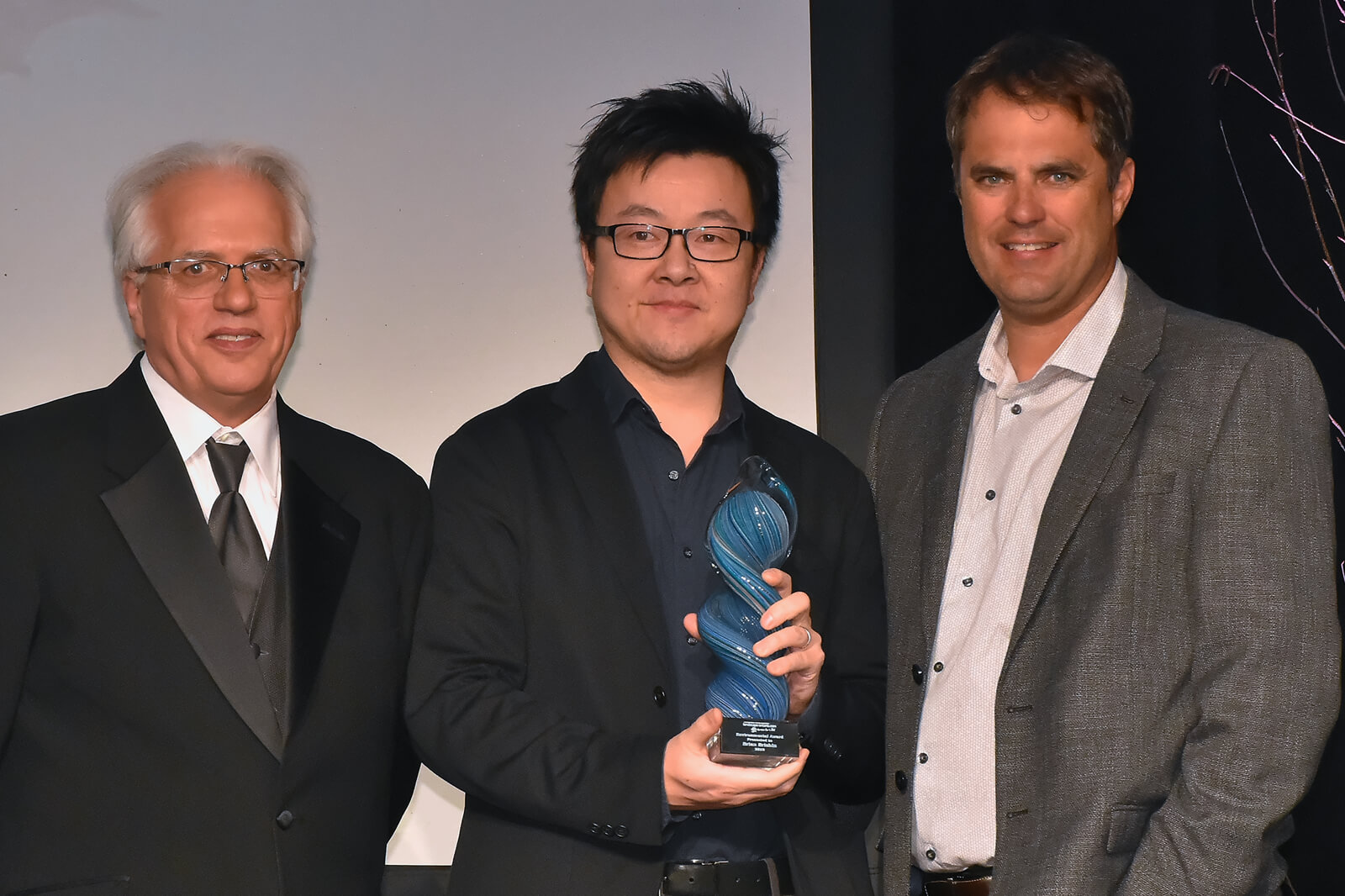 three men on stage. middle holding an award