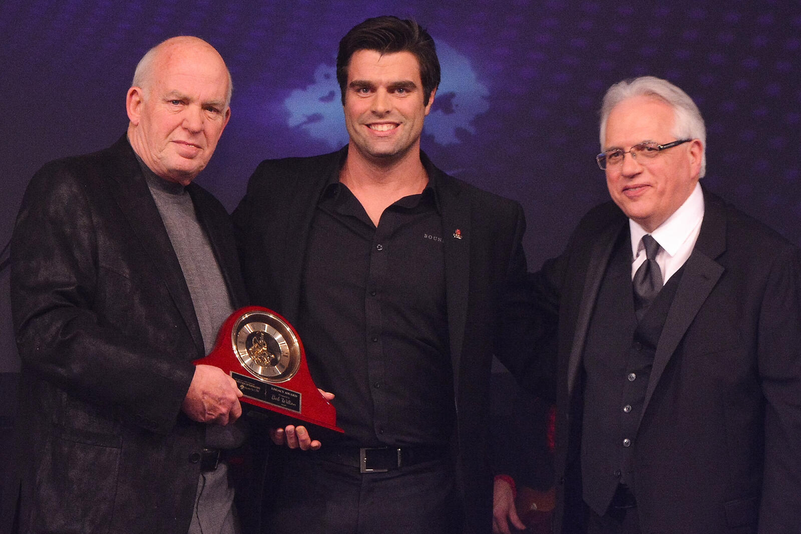 three men on stage, one holding an awrd