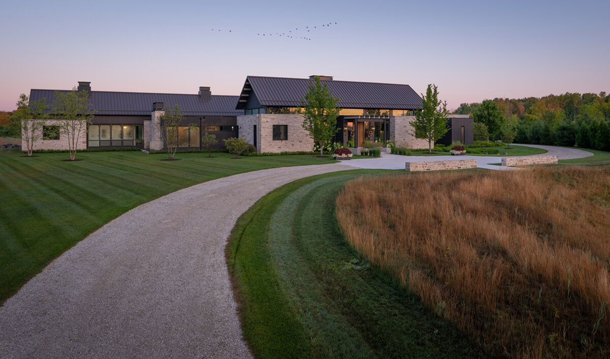 a long stone home with a metal roof at the end of a winding gravel road and perfect green lawn