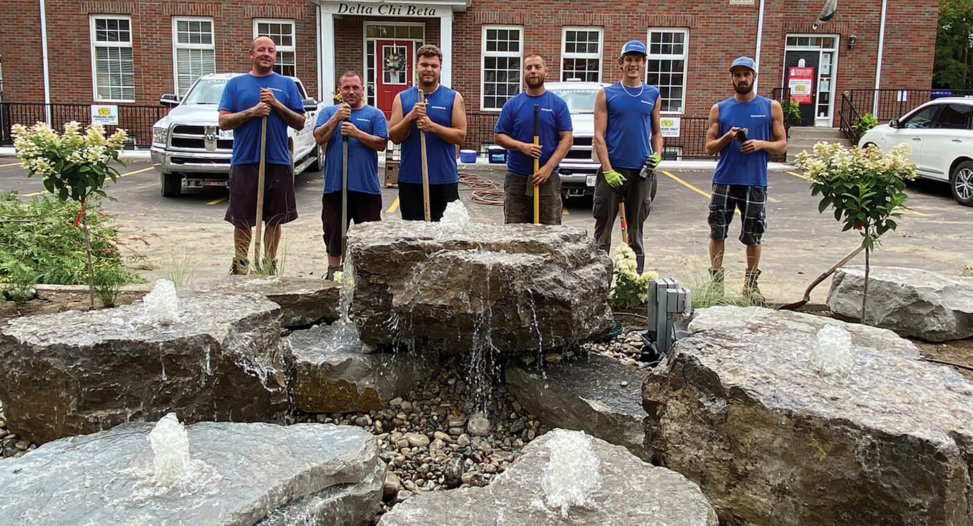 group of six workers in blue shirts stopping work for a photo in frong of a natural stone waterfeature in a parking lot