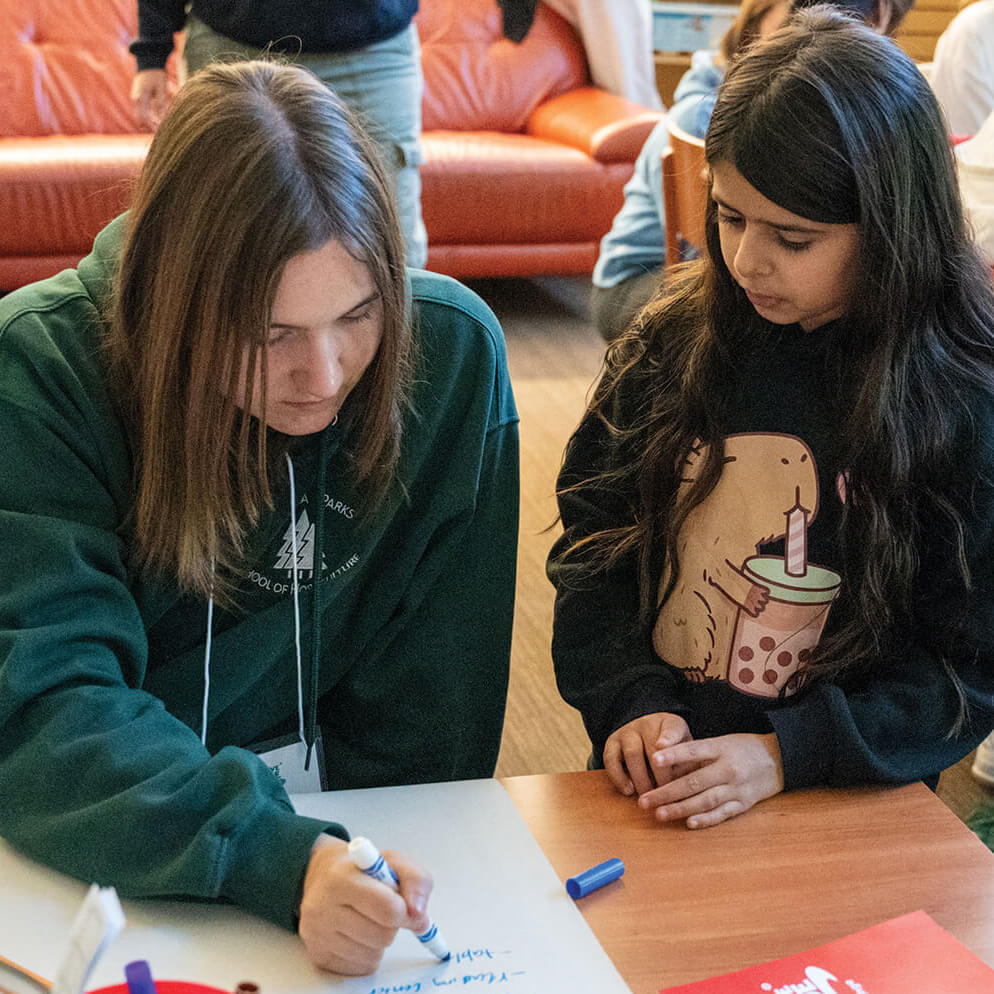 a young person colouring at a table while a younger person watches