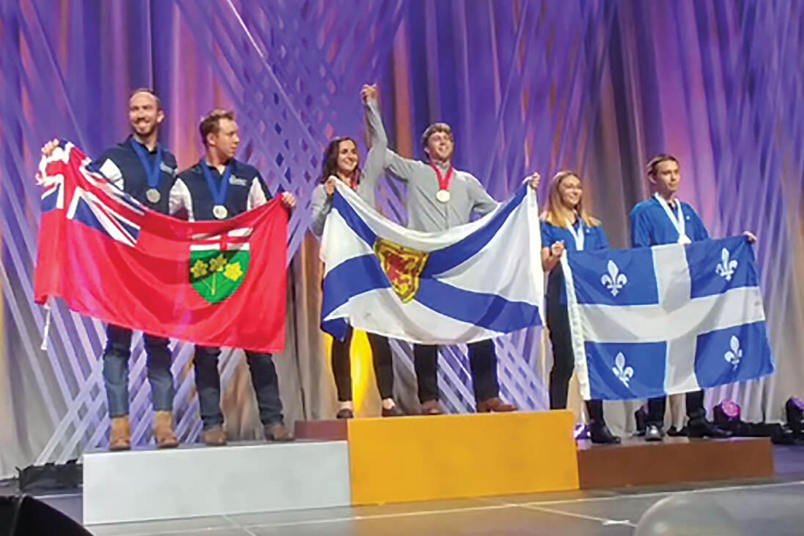 Algonquin College’s Blaise Mombourquett (far left) and Thomas Hawley hold the Ontario flag atop the silver podium at Skills Canada 2018 in Edmonton, Alta.